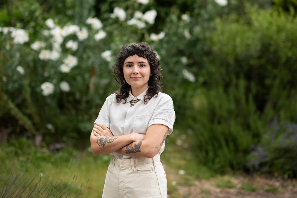 Amelia Rina in a white short sleeve button down shirt and off white pants standing in front of a background of lush plants.