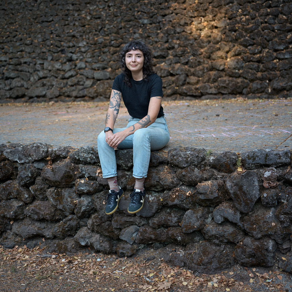 Amelia Rina in a black t-shirt and light wash jeans sits on a wall made of medium-sized gray rocks covered in brown moss that supports a concrete path. Behind her, there is another tall wall made of the same rocks.